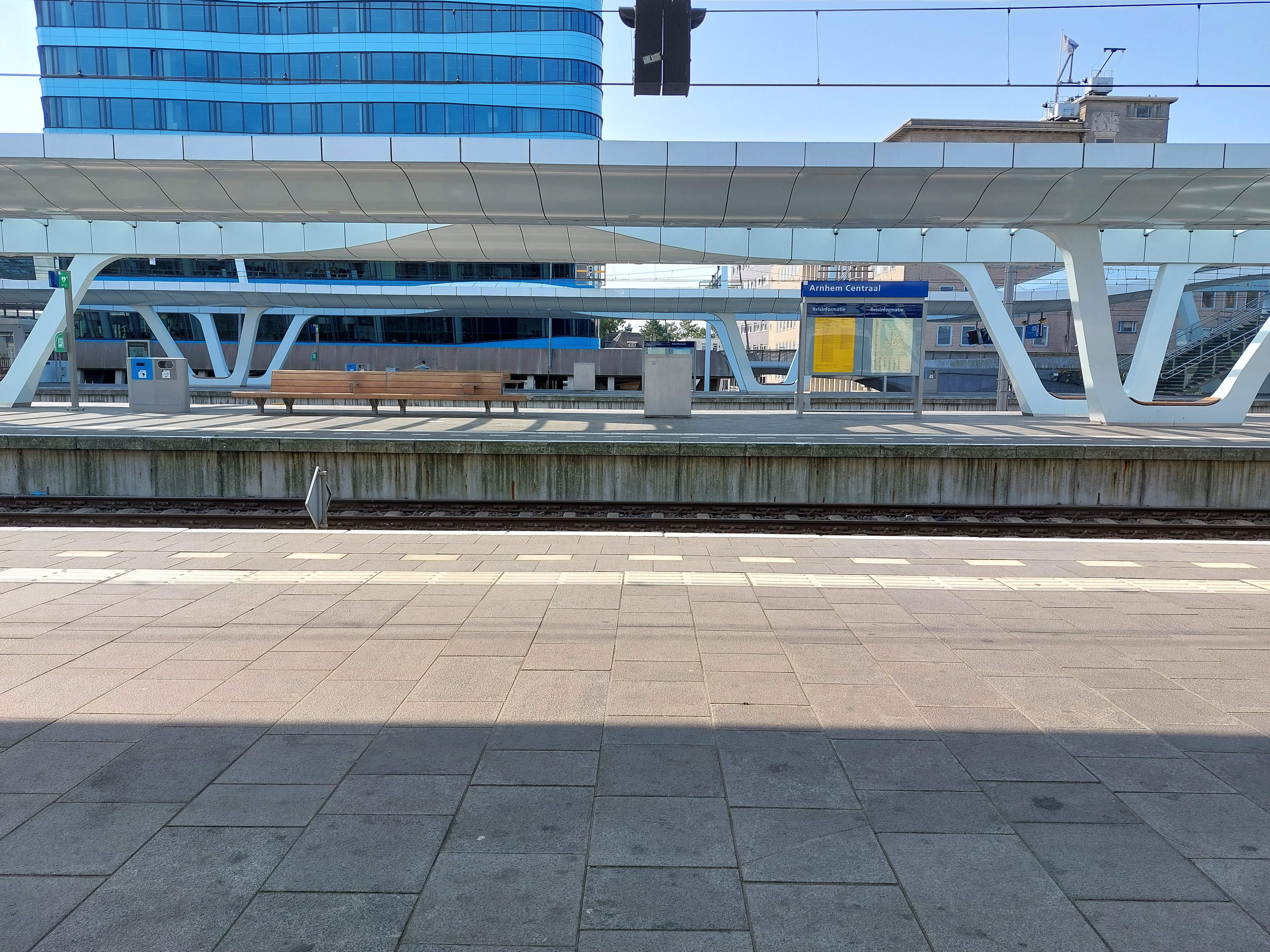 Foto van een perron. Op het perron staat onder andere een bankje en een bord "Arnhem Centraal" met reisinformatie. Op de achtergrond is een kantoorgebouw te zien.